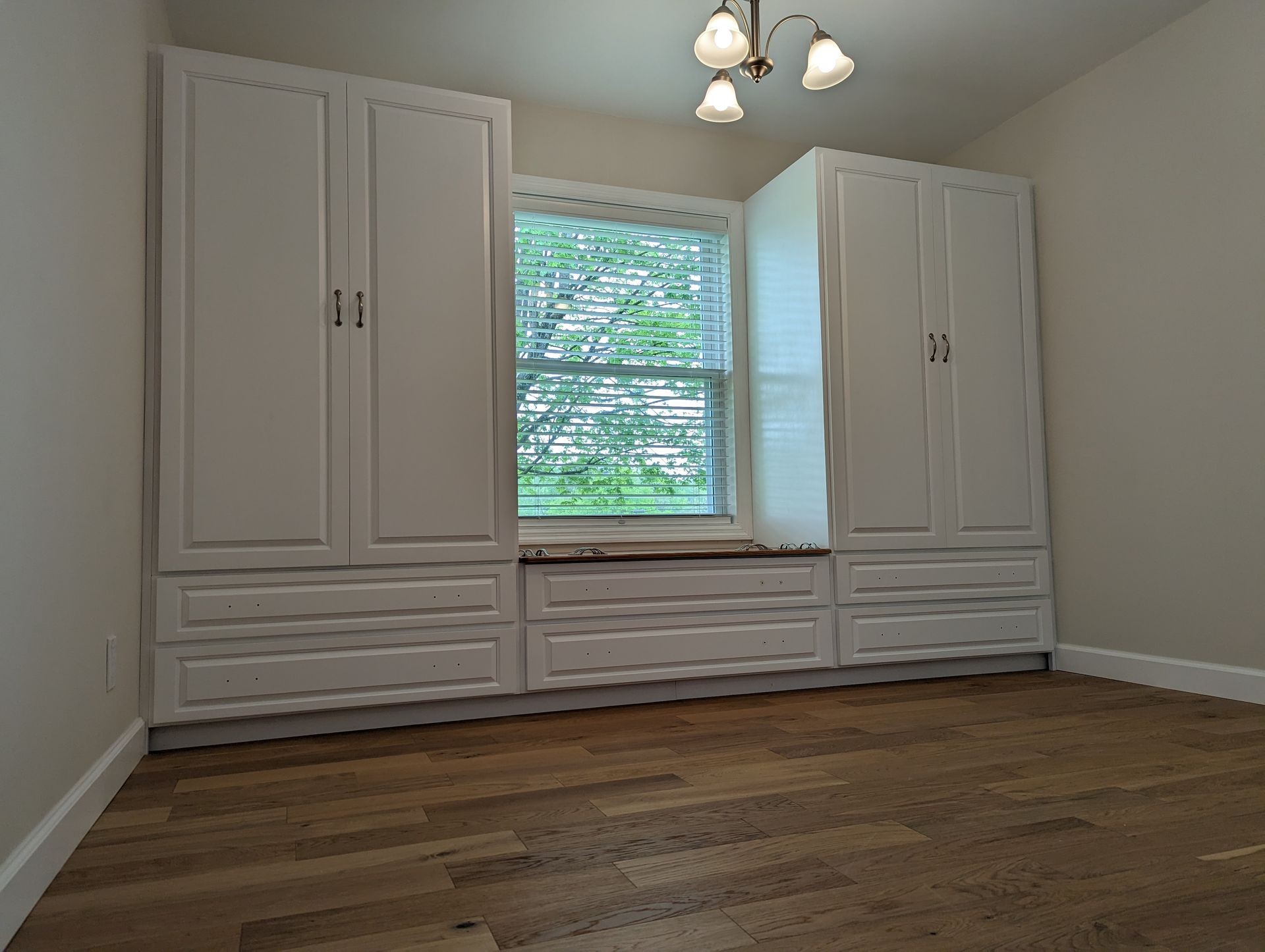 Empty room with white cabinets and a window seat, featuring a wooden floor and ceiling light fixture.