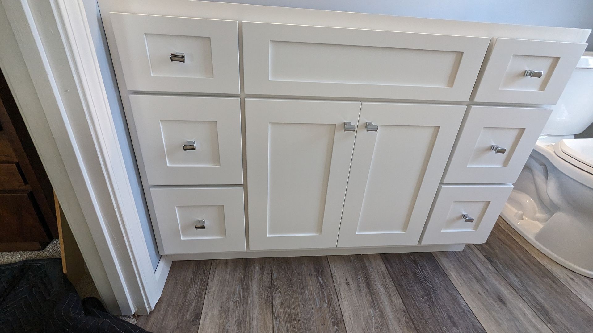 White bathroom vanity with six drawers and two cabinet doors, featuring metallic handles, on wood flooring.